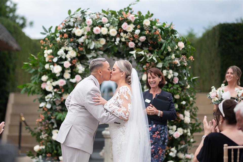 Shortmead House wedding flowers. Bedfordshire outdoor wedding venue ceremony with full flower arch and couple kissing in front of it